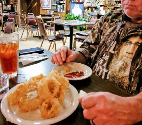 A person in a camouflage shirt holds a plate of fried onion rings with dipping sauce in a casual dining setting.