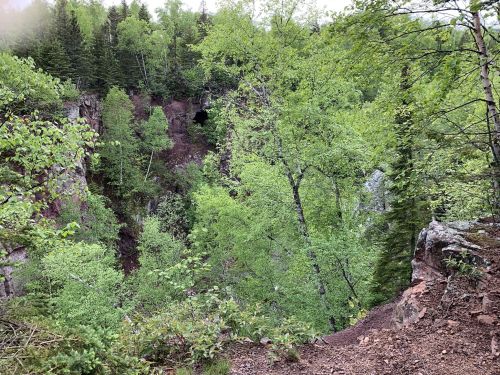 Lush green forest surrounds a rocky cliff with a cave entrance, creating a serene natural landscape.