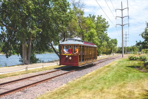 A vintage trolley travels along tracks beside a river, surrounded by trees and grassy areas on a sunny day.
