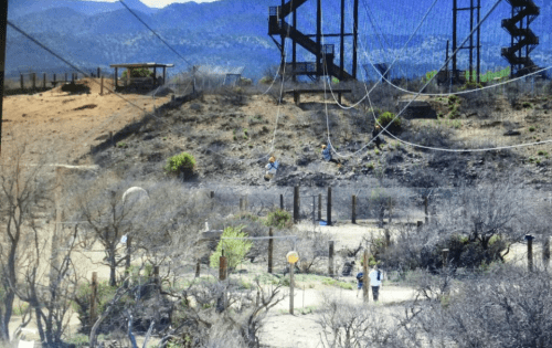 A landscape with dry terrain, a zip line, and people walking near structures against a mountainous backdrop.
