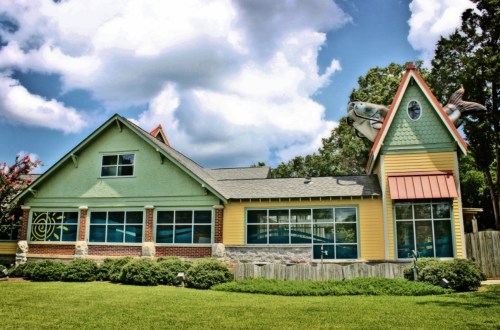 Colorful, whimsical building with a large fish sculpture on the roof, surrounded by green grass and trees under a blue sky.