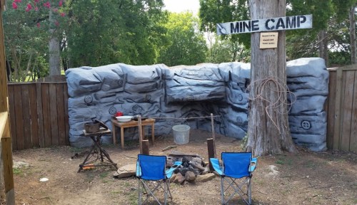 A rustic mine camp setup with a stone backdrop, wooden table, chairs, and tools in a wooded area.