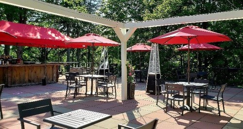 Outdoor patio with red umbrellas, tables, and chairs surrounded by greenery. A bar area is visible in the background.