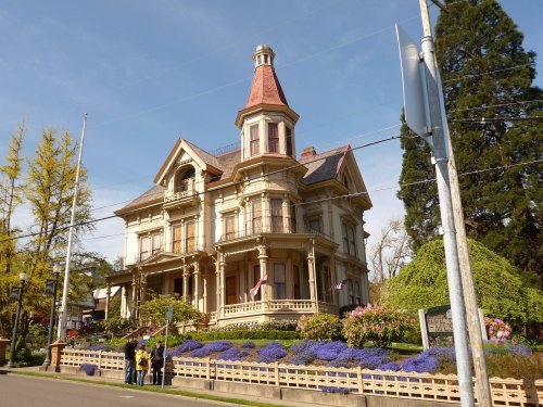 A large, ornate Victorian house with a turret, surrounded by colorful flowers and trees, with people in front.