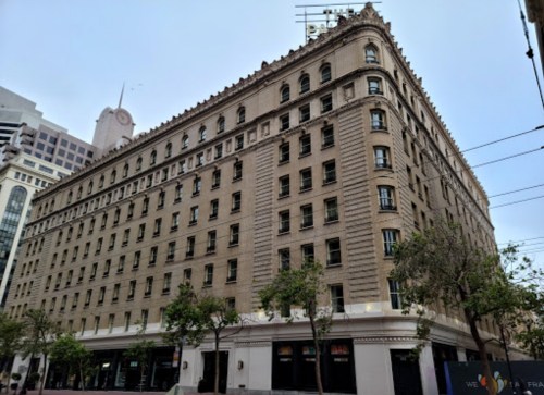 Historic building with a classic facade, featuring multiple windows and trees in the foreground, set against a cloudy sky.