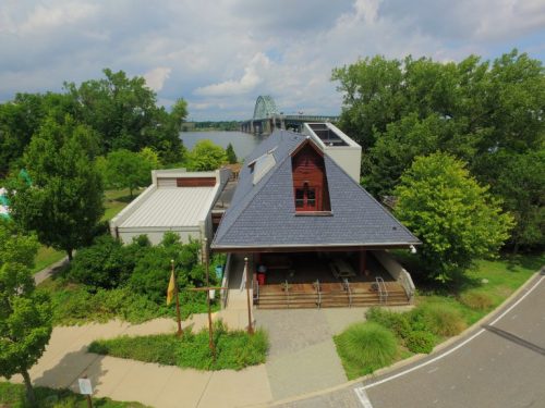 A modern building with a sloped roof surrounded by greenery, near a river and bridge under a cloudy sky.