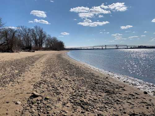 A sandy beach with pebbles, trees in the background, and a bridge over a sparkling river under a clear blue sky.