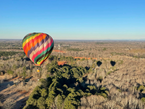 A colorful hot air balloon floats above a wooded landscape, casting a shadow on the trees below.