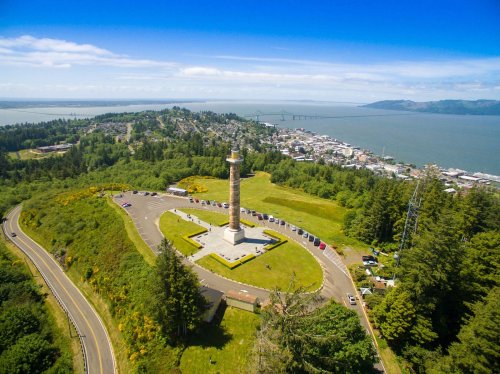Aerial view of a tall monument surrounded by greenery, overlooking a coastal town and waterway.