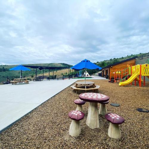 Playground area with colorful equipment, picnic tables, and umbrellas, surrounded by gravel and green hills in the background.