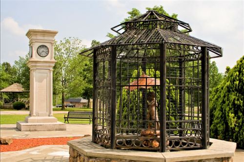 A decorative gazebo with a clock tower nearby, surrounded by greenery and a bench in a park setting.