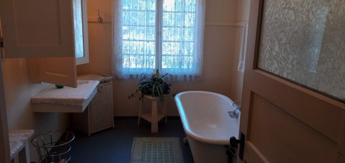 Bright bathroom with a bathtub, window with sheer curtains, and a potted plant, featuring light-colored walls and flooring.