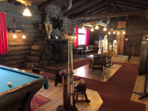 Interior of a rustic log cabin with a pool table, fireplace, and vintage decor, featuring mannequins in period clothing.