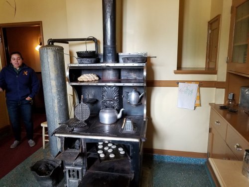 A vintage kitchen with a large stove, kettle, and baked goods, featuring a person standing nearby.