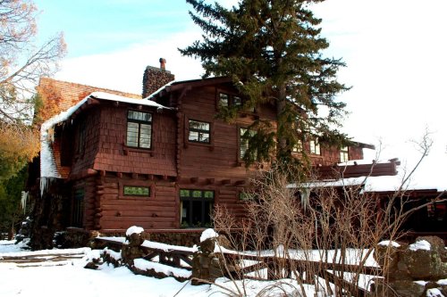 A large, rustic wooden house surrounded by snow, featuring multiple windows and a tall evergreen tree nearby.