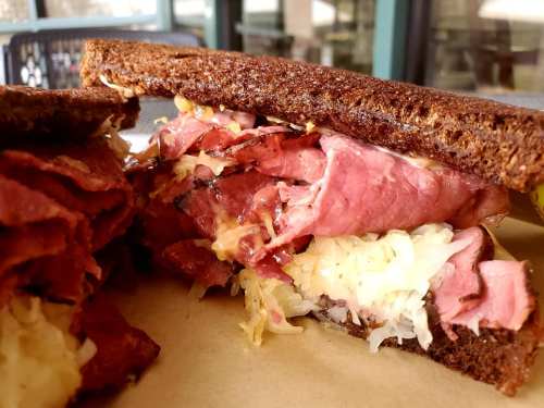 Close-up of a hearty sandwich with layers of pastrami, sauerkraut, and melted cheese on dark rye bread.