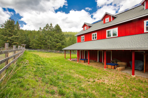 A red barn with a gray roof, surrounded by green grass and trees, under a blue sky with fluffy white clouds.