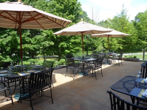 Outdoor dining area with black metal tables and chairs, shaded by striped umbrellas, surrounded by greenery.