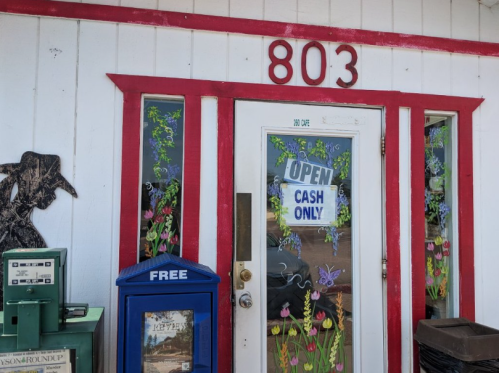 A storefront with a red-trimmed door, colorful floral decorations, and signs indicating "Open" and "Cash Only."