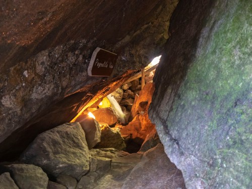 A narrow cave passage with rocks and a wooden sign reading "Pygmy's Hole" illuminated by soft light.