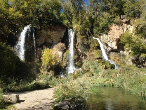 A scenic view of two waterfalls cascading down rocky cliffs, surrounded by lush greenery and a clear stream.