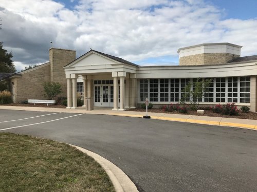 A modern building with large windows, stone accents, and a landscaped entrance under a cloudy sky.