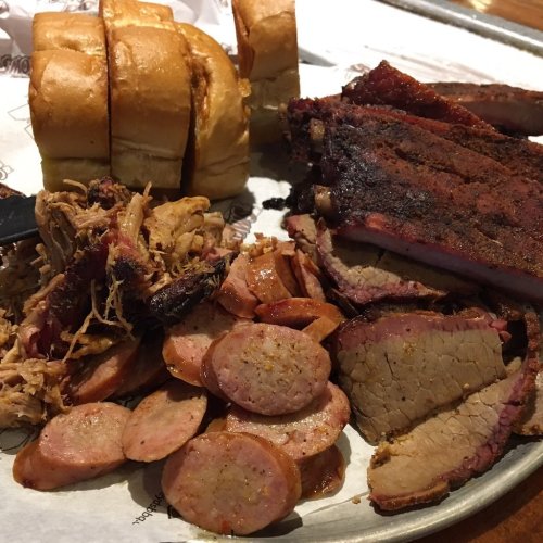 A platter of barbecue featuring sliced brisket, pulled pork, sausage, and soft bread rolls.