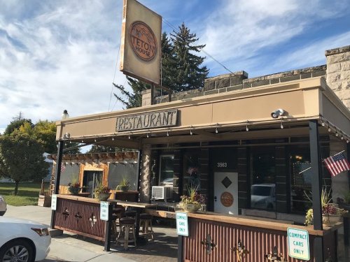 Exterior of Teton House restaurant with a sign, outdoor seating, and flags, set against a blue sky.