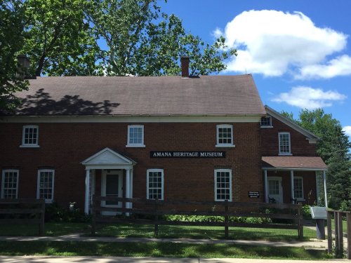 Amana Heritage Museum building with a brick facade, surrounded by trees and a blue sky with clouds.