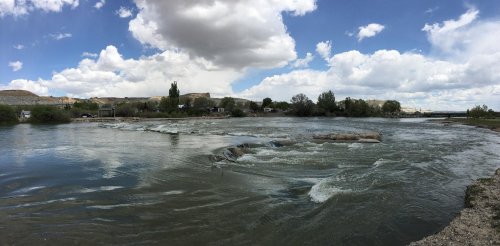 A serene river flows under a cloudy sky, with trees lining the banks and gentle rapids visible in the water.