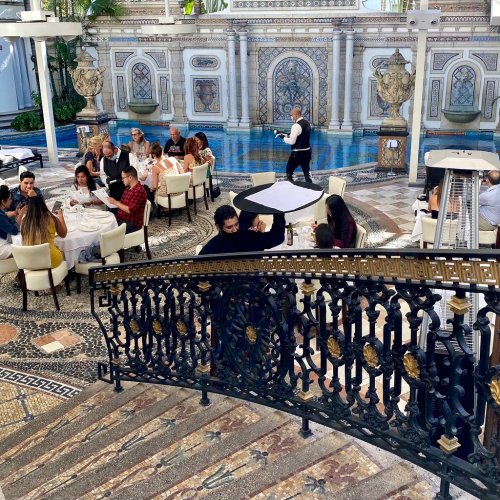 A bustling restaurant scene with diners at tables, a waiter serving, and an ornate backdrop featuring a pool and architecture.