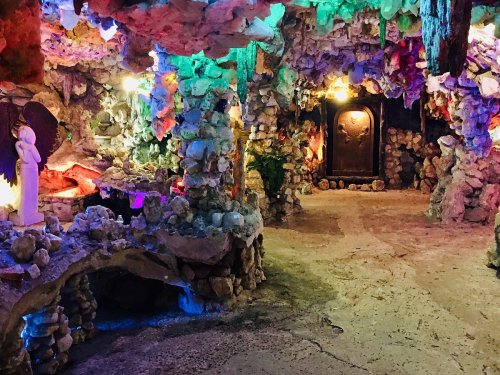 Colorful cave interior with stone formations, illuminated by vibrant lights, featuring sculptures and a dark doorway.