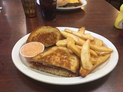 A plate with a grilled sandwich, fries, and a small cup of dipping sauce on a wooden table.