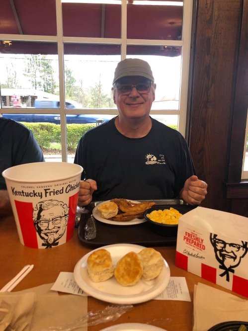 A smiling man sits at a table with a bucket of fried chicken, biscuits, and macaroni and cheese in a restaurant.