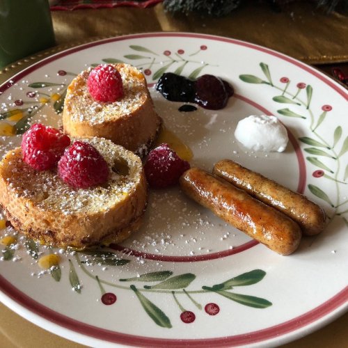 A plate of French toast topped with raspberries, syrup, and sausage links, garnished with powdered sugar.
