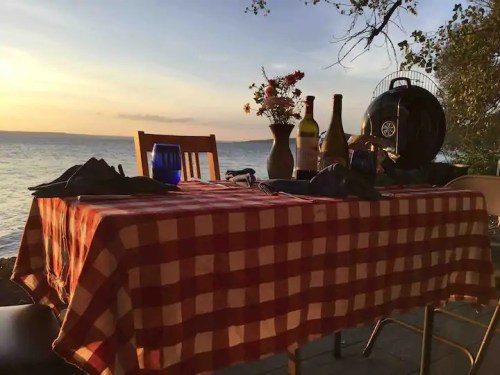 A sunset view of a table set with a checkered tablecloth, wine bottles, and flowers by the water's edge.
