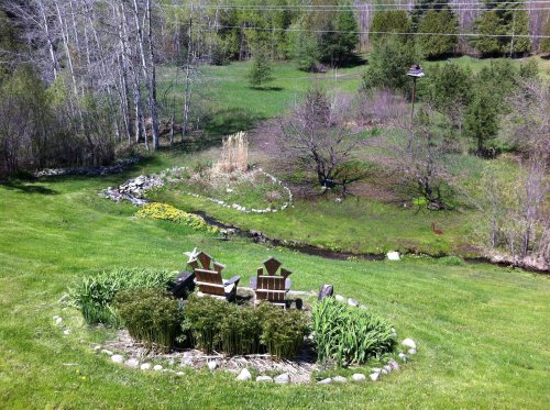 A serene garden scene with two chairs, a stream, and lush greenery, surrounded by trees and blooming flowers.