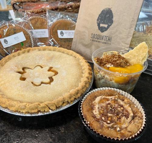 A variety of baked goods including a pie, dessert cup, and muffins, displayed on a countertop with packaging in the background.