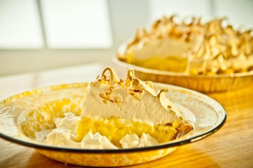 A slice of lemon meringue pie on a plate, with a whole pie in the background, topped with whipped cream and toasted meringue.
