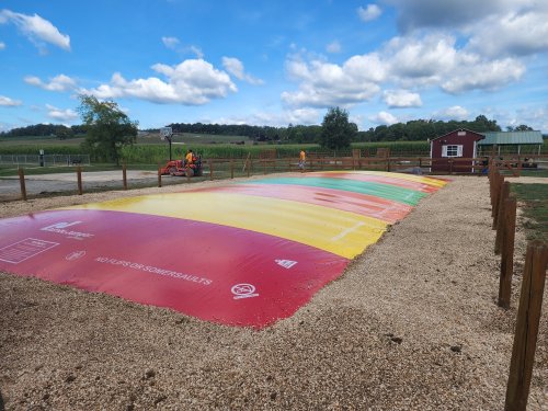 A colorful inflatable jumping pad in a gravel area, surrounded by wooden fencing and green fields under a blue sky.