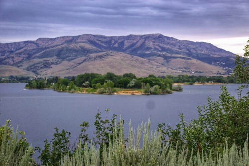A serene lake surrounded by mountains, featuring a small island with lush greenery under a cloudy sky.