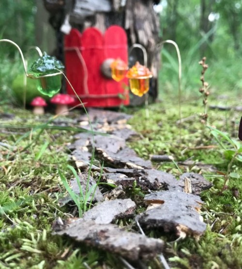 A whimsical scene featuring colorful glass flowers and a small red door set among moss and bark in a forest.