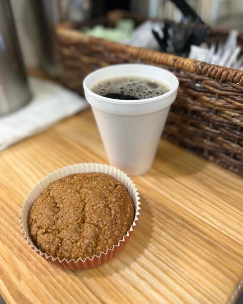 A warm muffin in a paper liner next to a cup of black coffee on a wooden surface.
