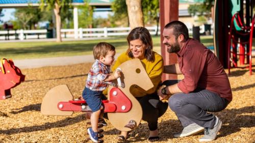 A family enjoys time at a playground, with a young boy playing on a rocking horse while his parents smile at him.