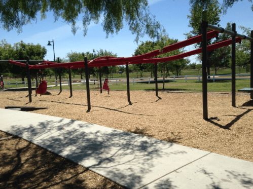 A playground with red climbing structures and a sandy area, surrounded by trees and open green space.