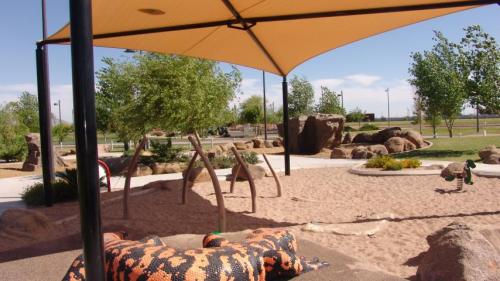 A playground with a sandy area, climbing structures, and a large, colorful animal sculpture under a shaded canopy.