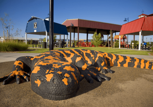 A large, colorful lizard sculpture with orange and black patterns, set in a park with playground structures in the background.