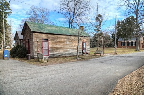 A weathered wooden building with a green roof, benches, and trees, set along a quiet road under a blue sky.
