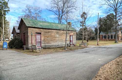 A weathered wooden building with a green roof, benches, and trees, set along a quiet road under a blue sky.