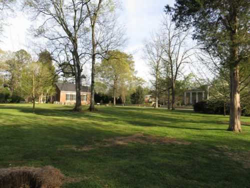 A spacious green lawn with trees, featuring a brick house and a distant building under a clear sky.